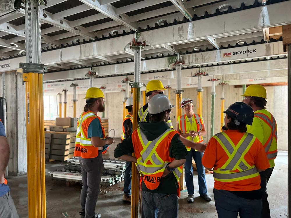 A group of young adults with hi-vis shirts and hard hats looking at a female with a hi-vis shirt and a hard hat in construction site