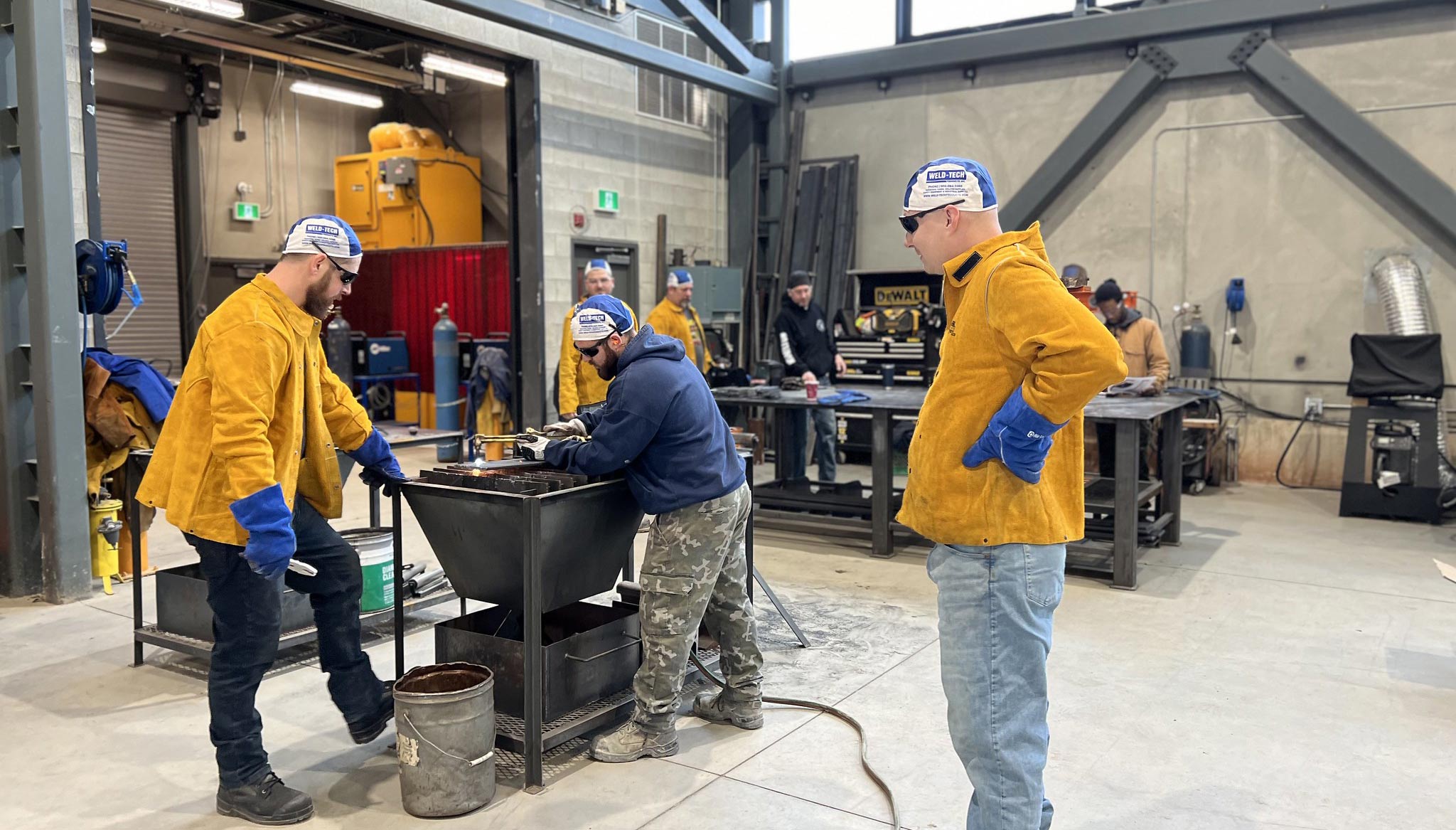 Three men in yellow jackets collaborate on a metal object, focused and engaged in their work