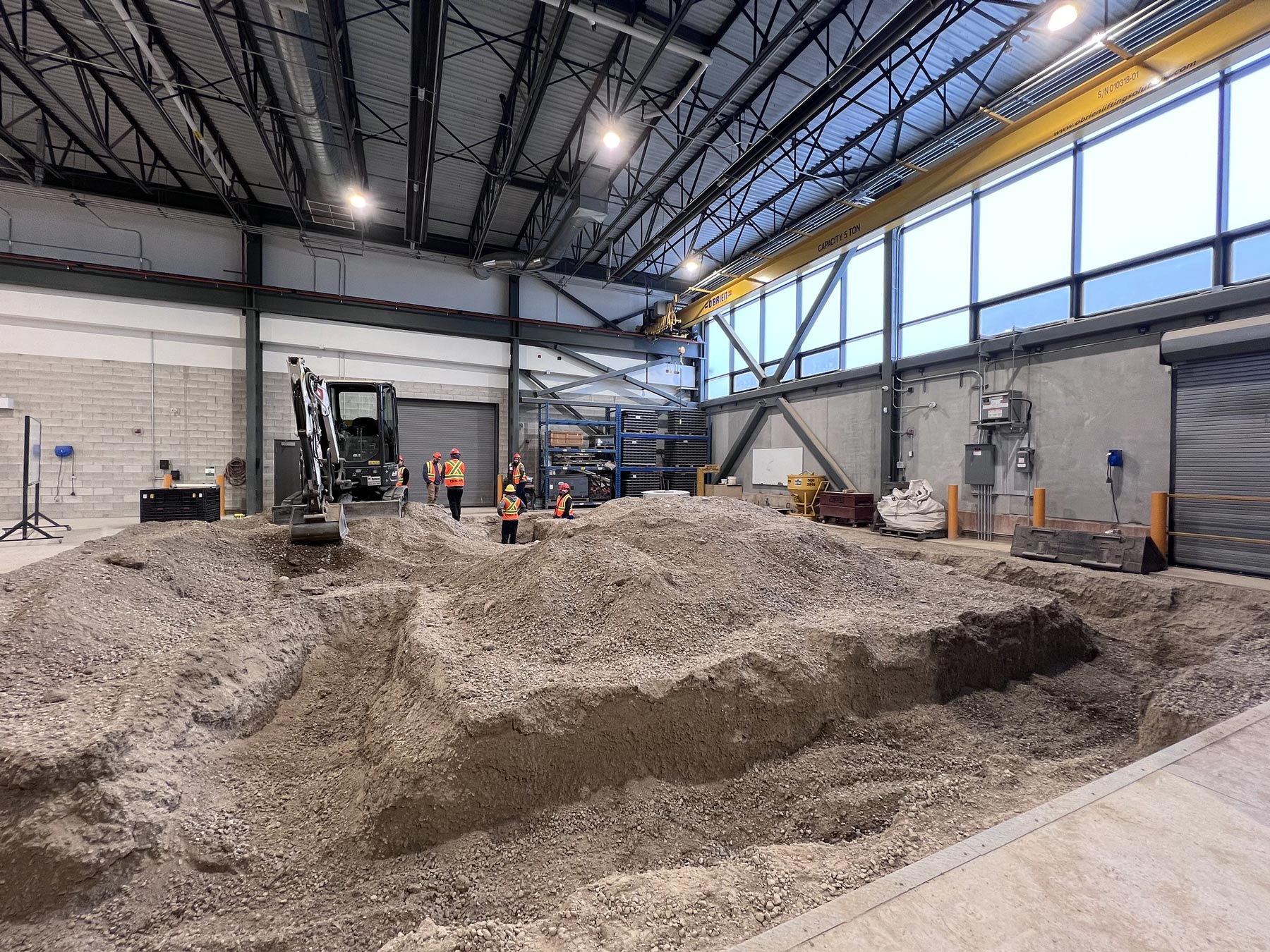 A big heap of soil in a warehouse, surrounded by concrete walls and illuminated by overhead lights. with construction workers interacting with each other