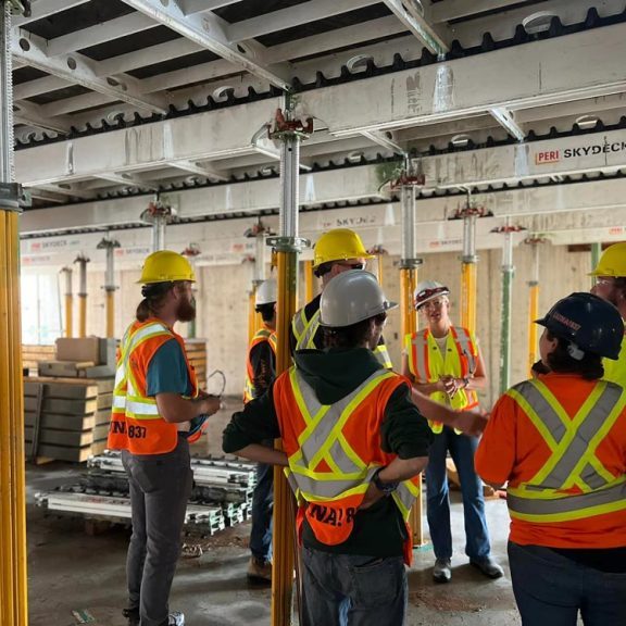 A group of young adults with hi-vis shirts and hard hats looking at a female with a hi-vis shirt and a hard hat in construction site