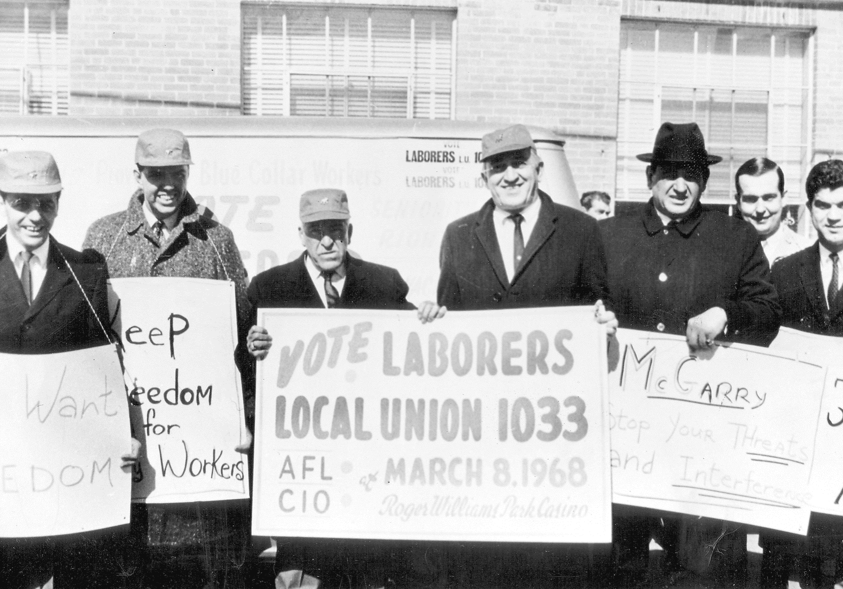 The black and white image features a group of men standing in front of a brick building, holding signs. The men are wearing coats and hats, indicative of cold weather. The central sign, held by two men, reads "VOTE LABORERS LOCAL UNION 1033 AFL CIO MARCH 8, 1968 Roger Williams Park Casino," suggesting a labor union gathering or protest. Additional signs read "Keep Freedom for Workers" and "McGarry Stop Your Threats and Interference." The men are smiling, appearing united in their cause. The brick wall behind them and partially visible windows suggest an urban setting.